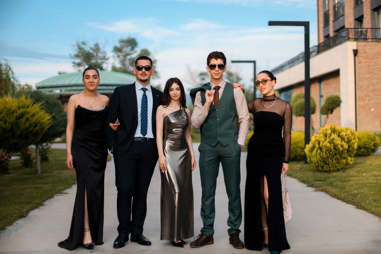 who-we-are A stylish group of five adults in formal attire posing outdoors on a sunny day.